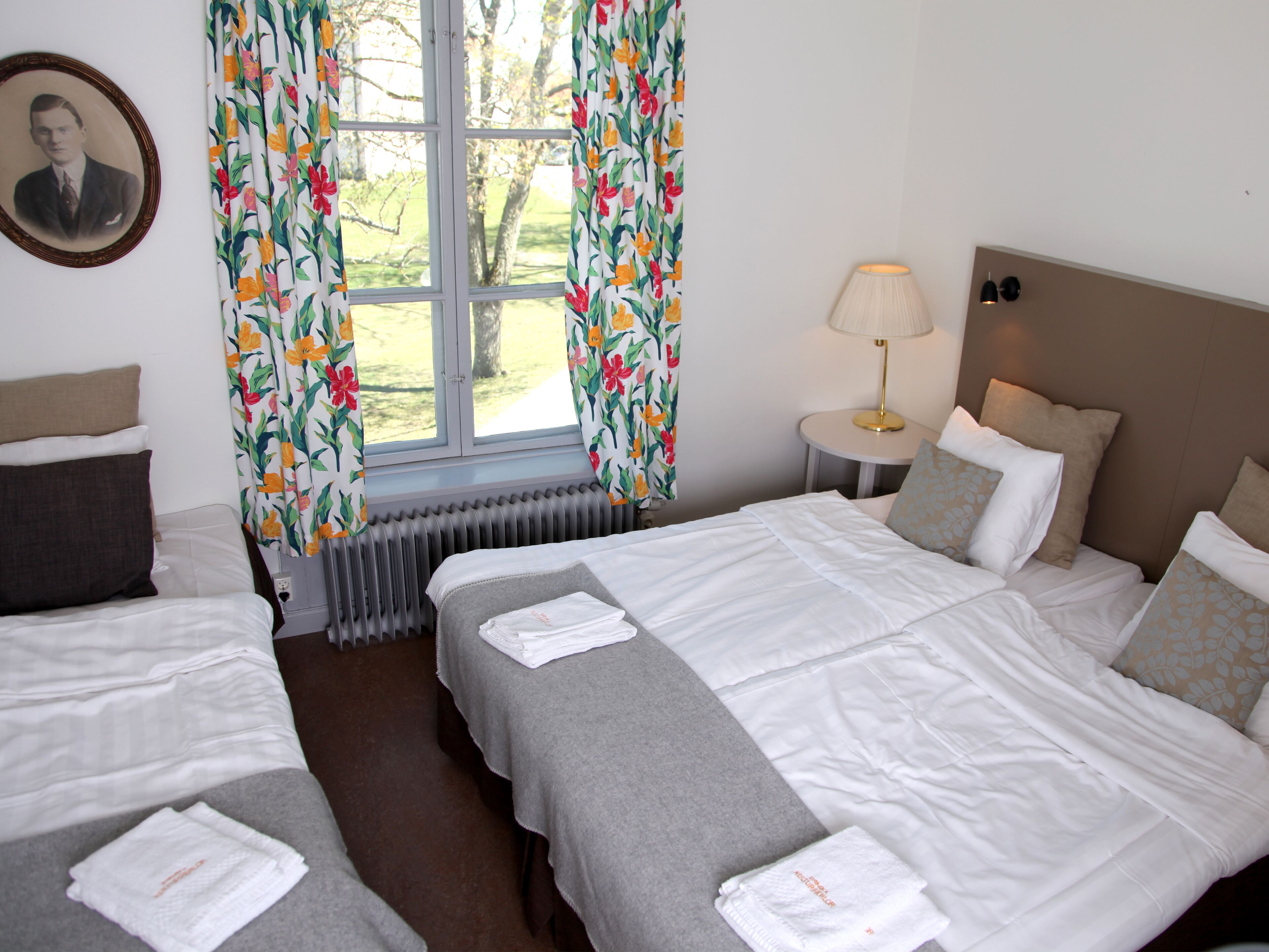 A hotel room with two beds, a radiator, a lamp, and a framed picture on the wall.