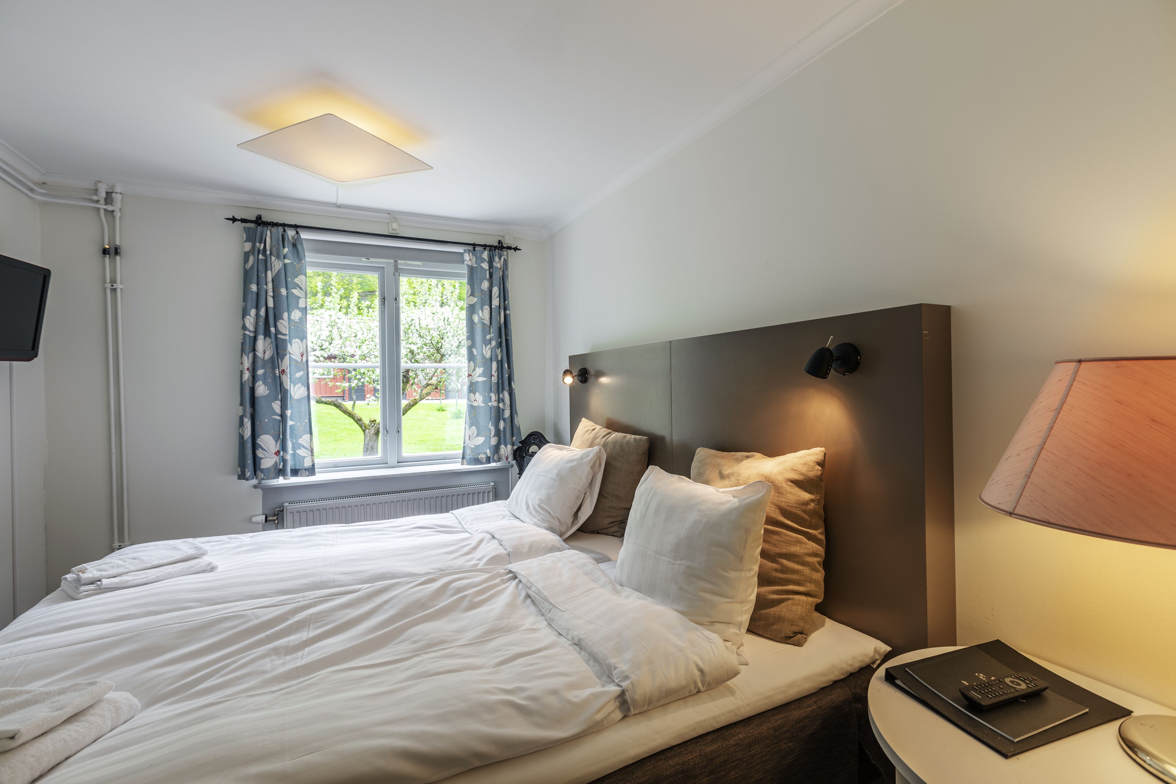 Una cama bien hecha con sábanas blancas, una mesita de noche con una lámpara y una ventana con cortinas.