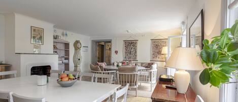 A dining area with a white table, chairs, and a bowl of fruit. There is a kitchen area in the background with a stove and cabinets.