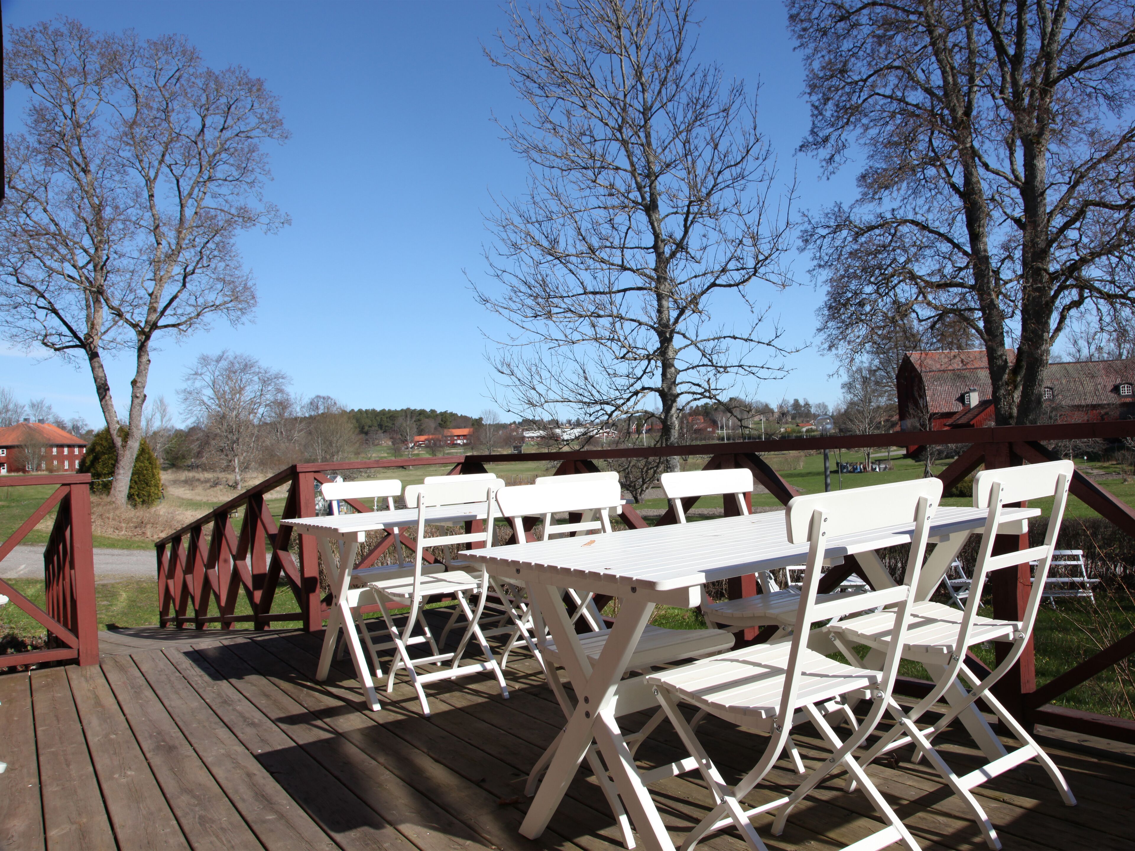 Una terraza de madera con mesas y sillas plegables blancas, con vistas a un paisaje rural con árboles y casas.