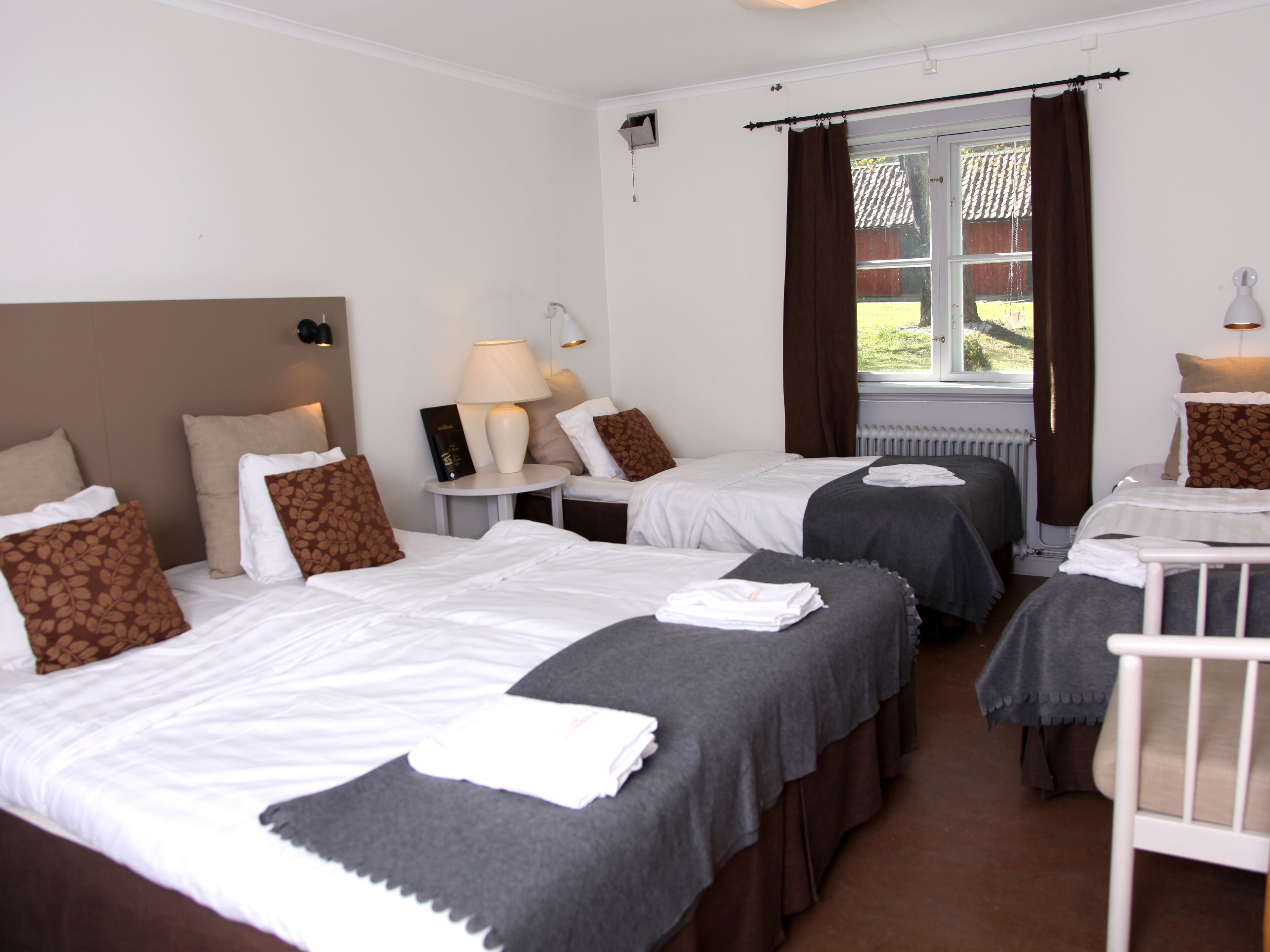 A hotel room with two beds, a window with curtains, a bedside table with a lamp, and a radiator.