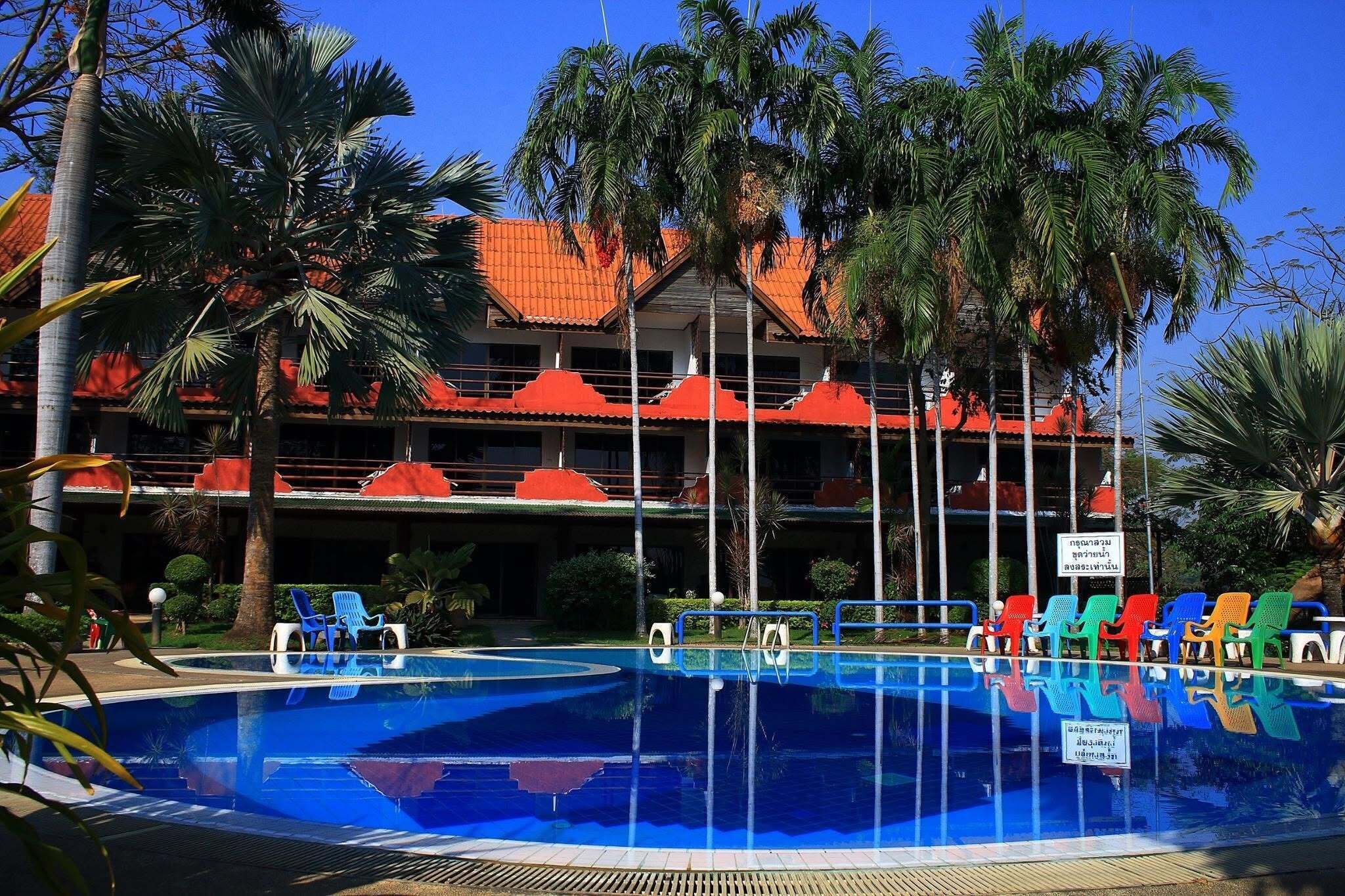 A swimming pool with multiple levels of balconies, surrounded by palm trees and a building with red-tiled roofs.