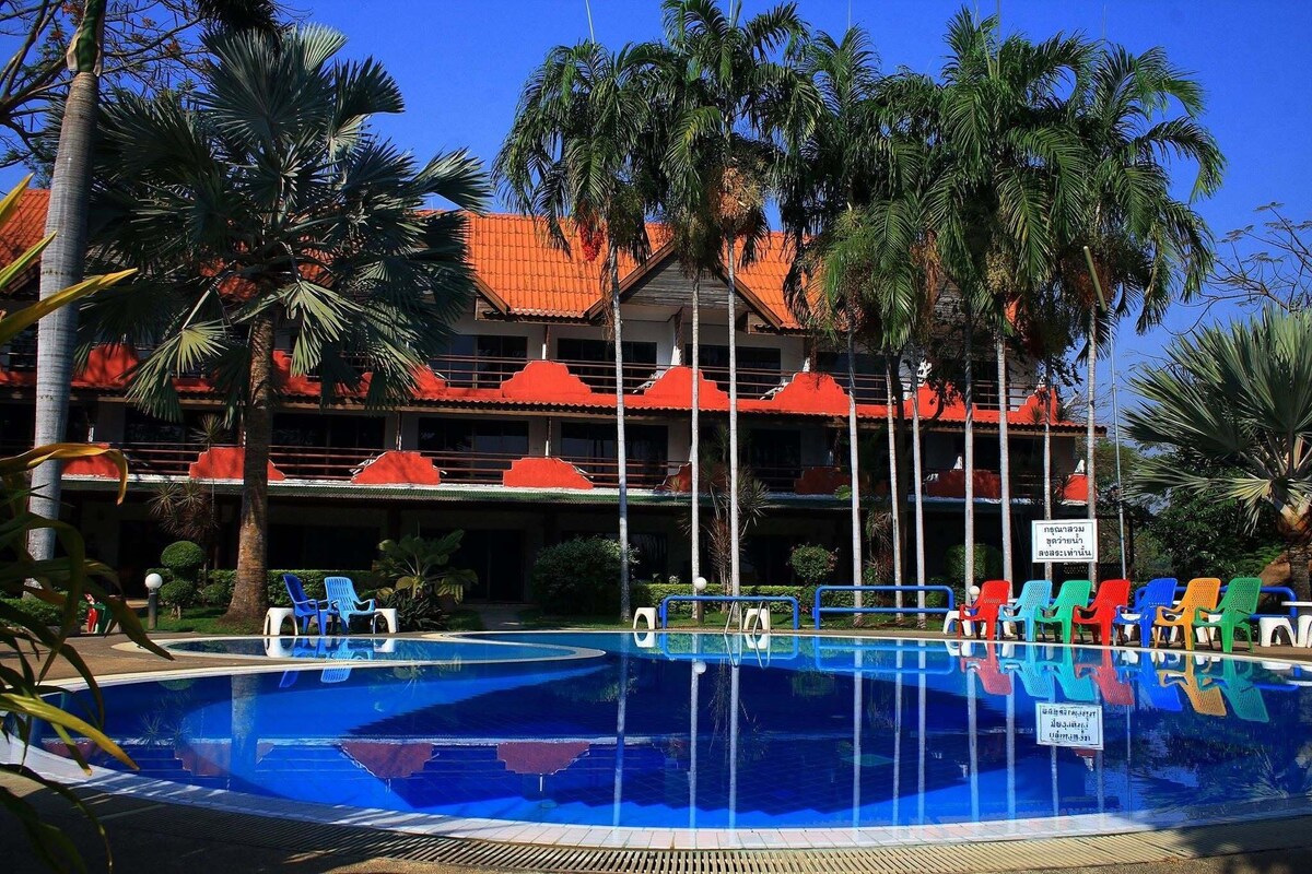 A swimming pool with multiple levels of balconies, surrounded by palm trees and a building with red-tiled roofs.