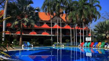 A swimming pool with multiple levels of balconies, surrounded by palm trees and a building with red-tiled roofs.