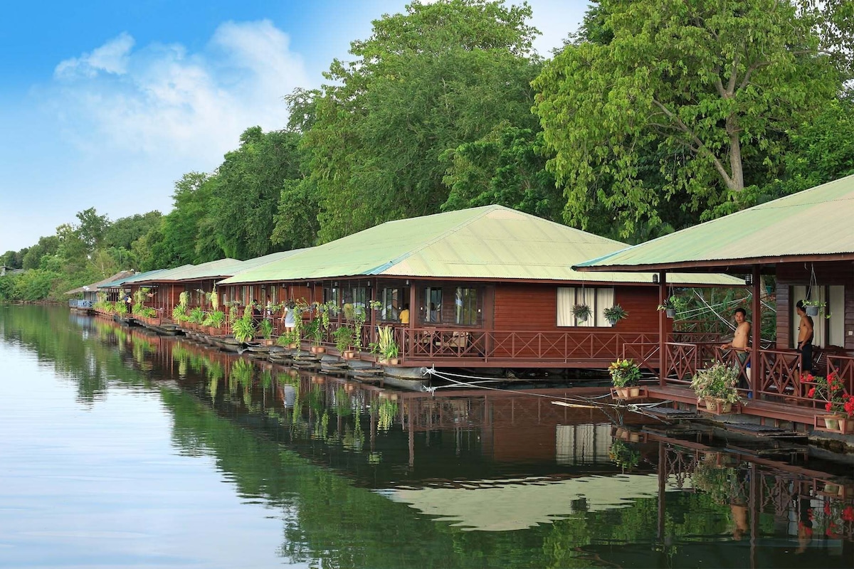 A row of wooden cabins with a river in the foreground and trees in the background.
