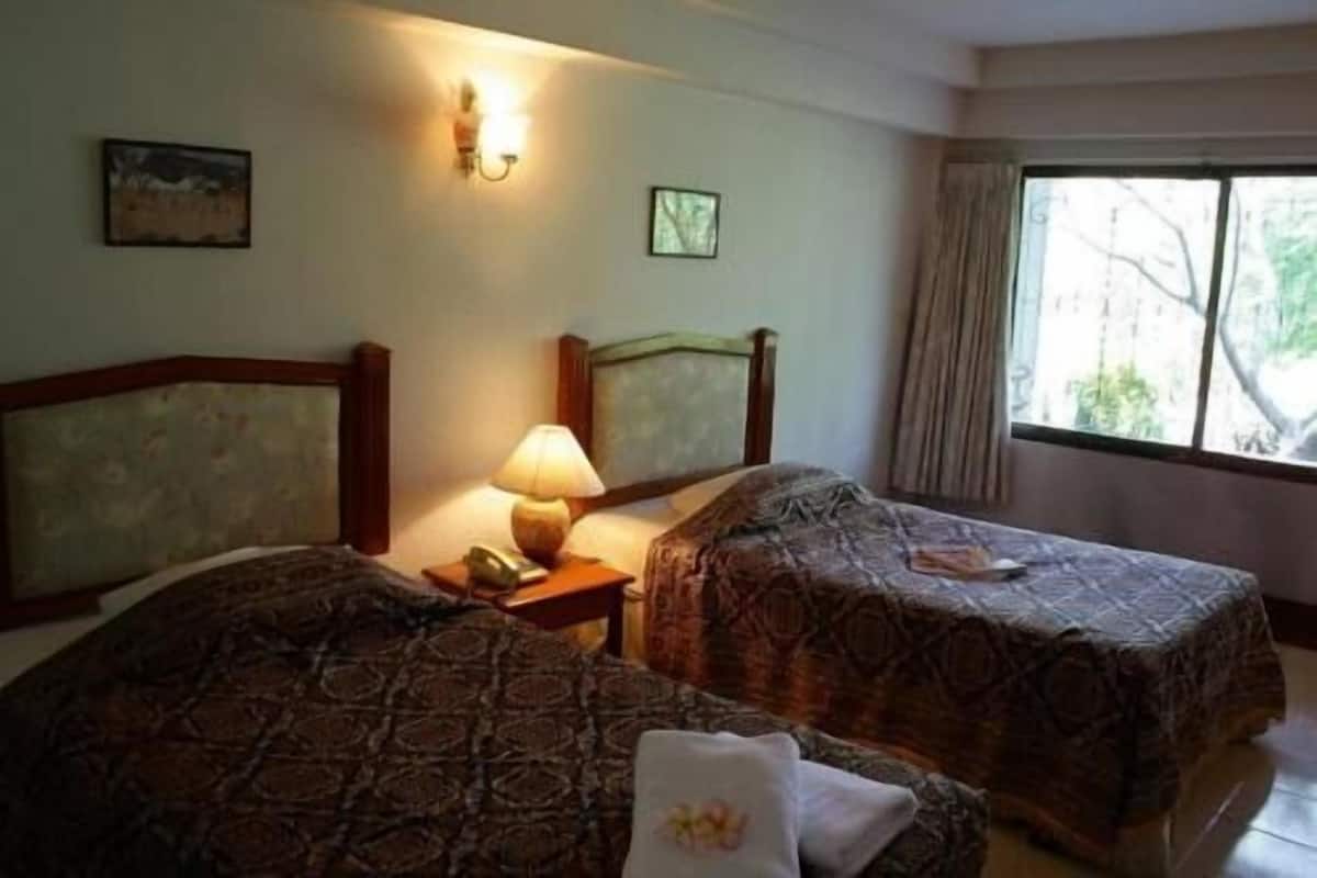 A hotel room with two beds, a nightstand with a lamp, a window with curtains, and a painting on the wall.