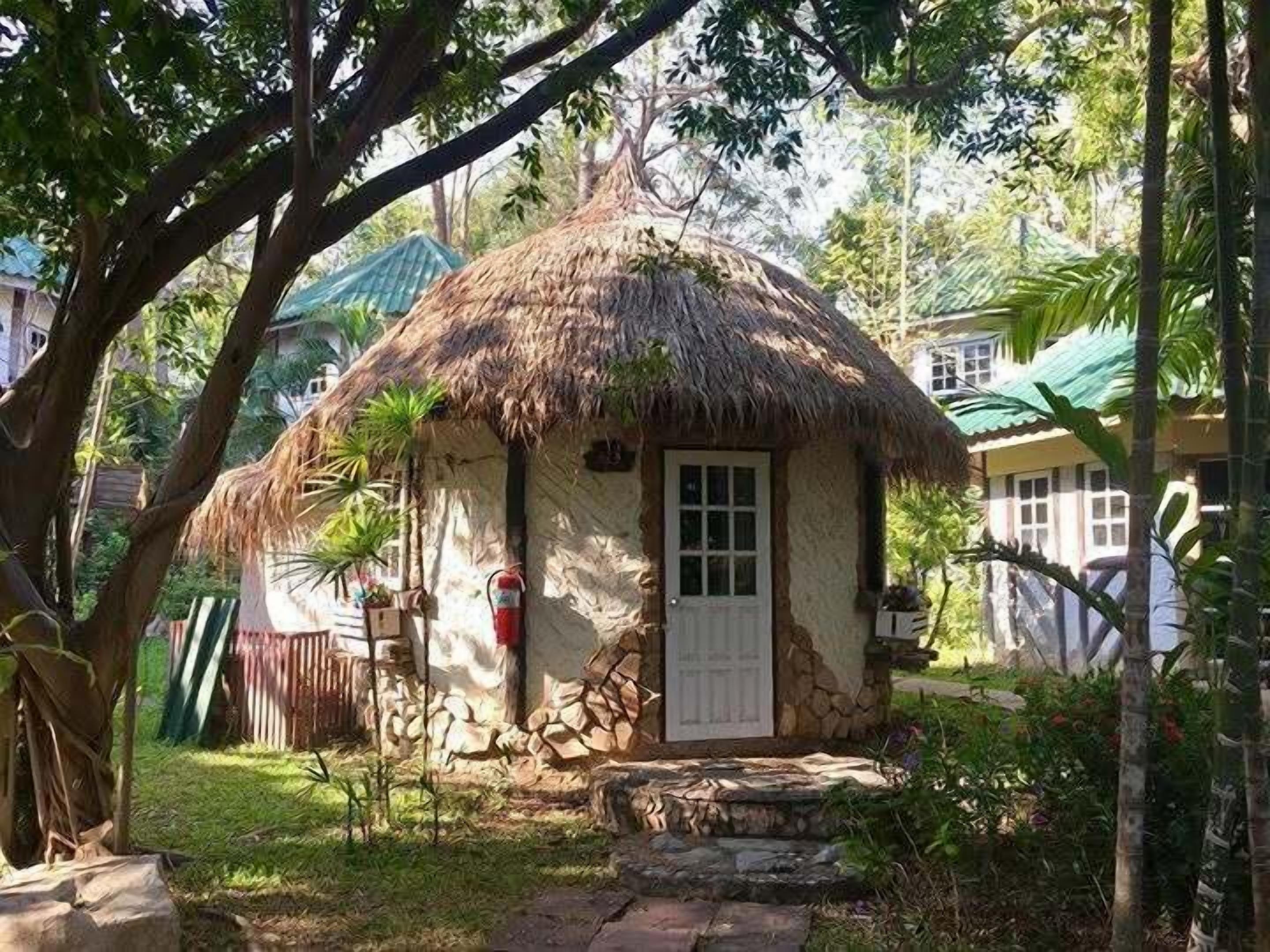 A thatched-roof hut with a door and windows, surrounded by greenery and a stone pathway.