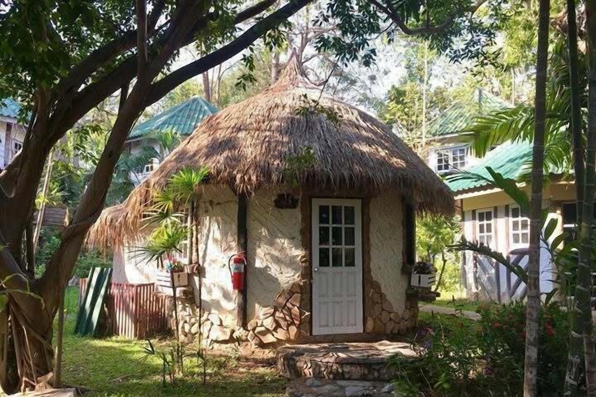 A thatched-roof hut with a door and windows, surrounded by greenery and a stone pathway.