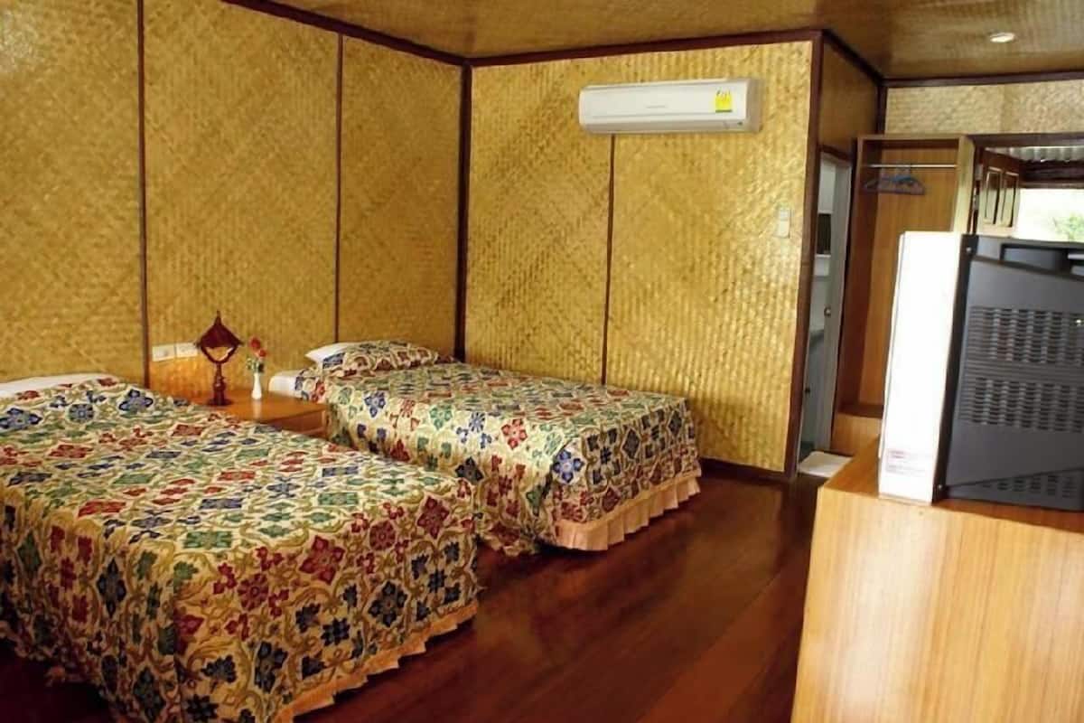 A room with two beds, a wooden floor, a wall-mounted air conditioner, and a small table with a lamp.