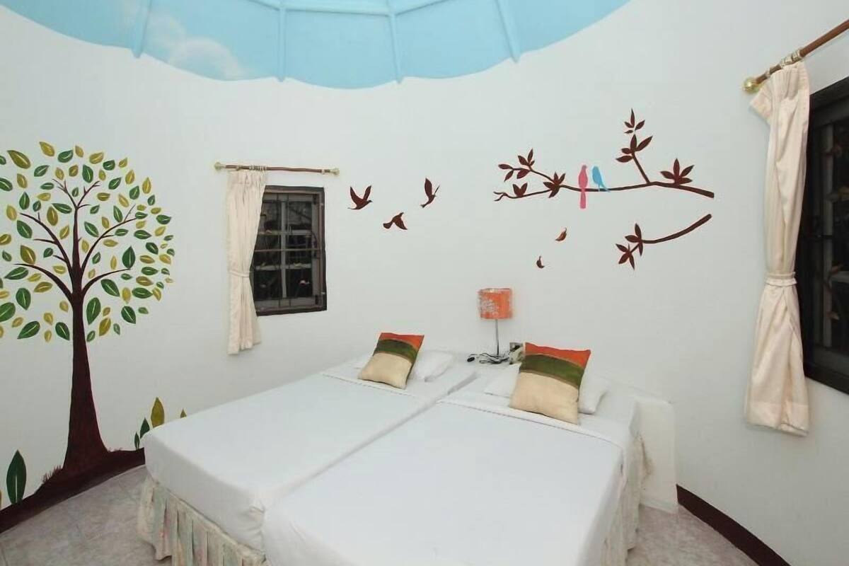 A room with two beds, a window, a tree mural, and a bird mural on the wall.