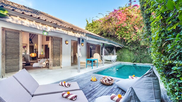A poolside area with lounge chairs and a small table, surrounded by lush greenery and a building with a tiled roof. - Villa 007 (Seminyak)