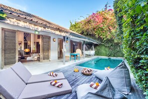 A poolside area with lounge chairs and a small table, surrounded by lush greenery and a building with a tiled roof. - Villa 007 (Seminyak)