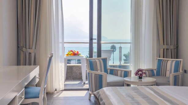 A hotel room with a bed, a desk, a chair, a small table with a vase of flowers, and a balcony with a view of the sea. - Hotel Bracera (Budva)