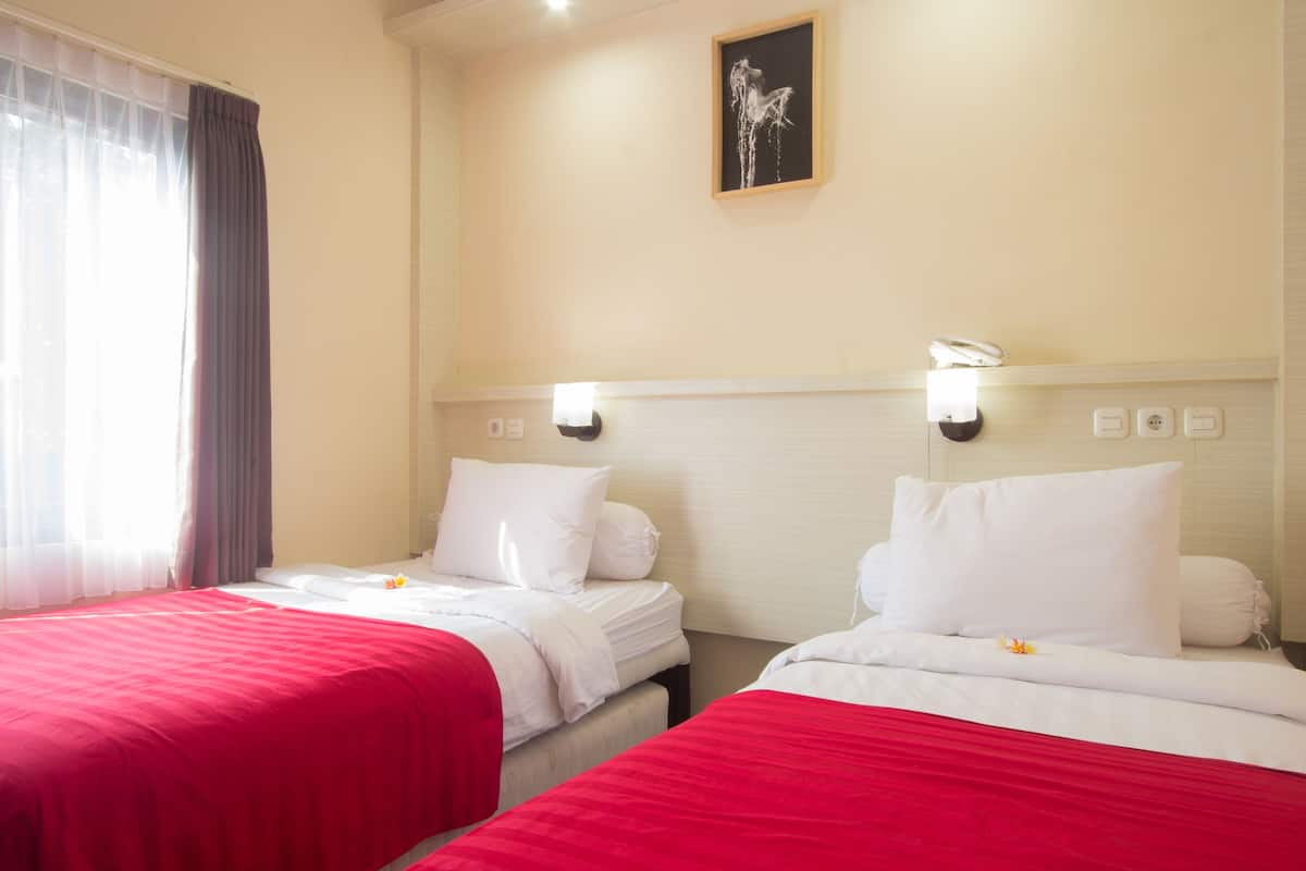A hotel room with two beds, each with red bedding and white pillows. There is a painting on the wall above the beds and a window with curtains on the left side.