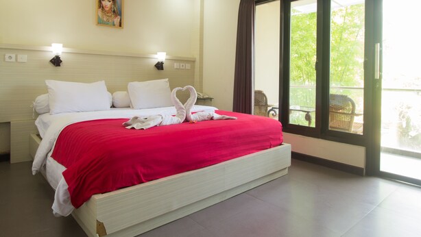 Une chambre d’hôtel avec un grand lit, une couverture rouge, des oreillers blancs, une serviette en forme de cœur, un tableau au mur et un balcon avec vue.