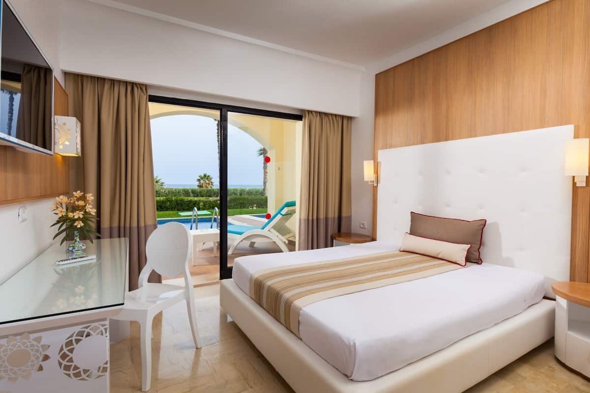 Habitación de hotel con cama, escritorio, silla, vista a la piscina y balcón.
