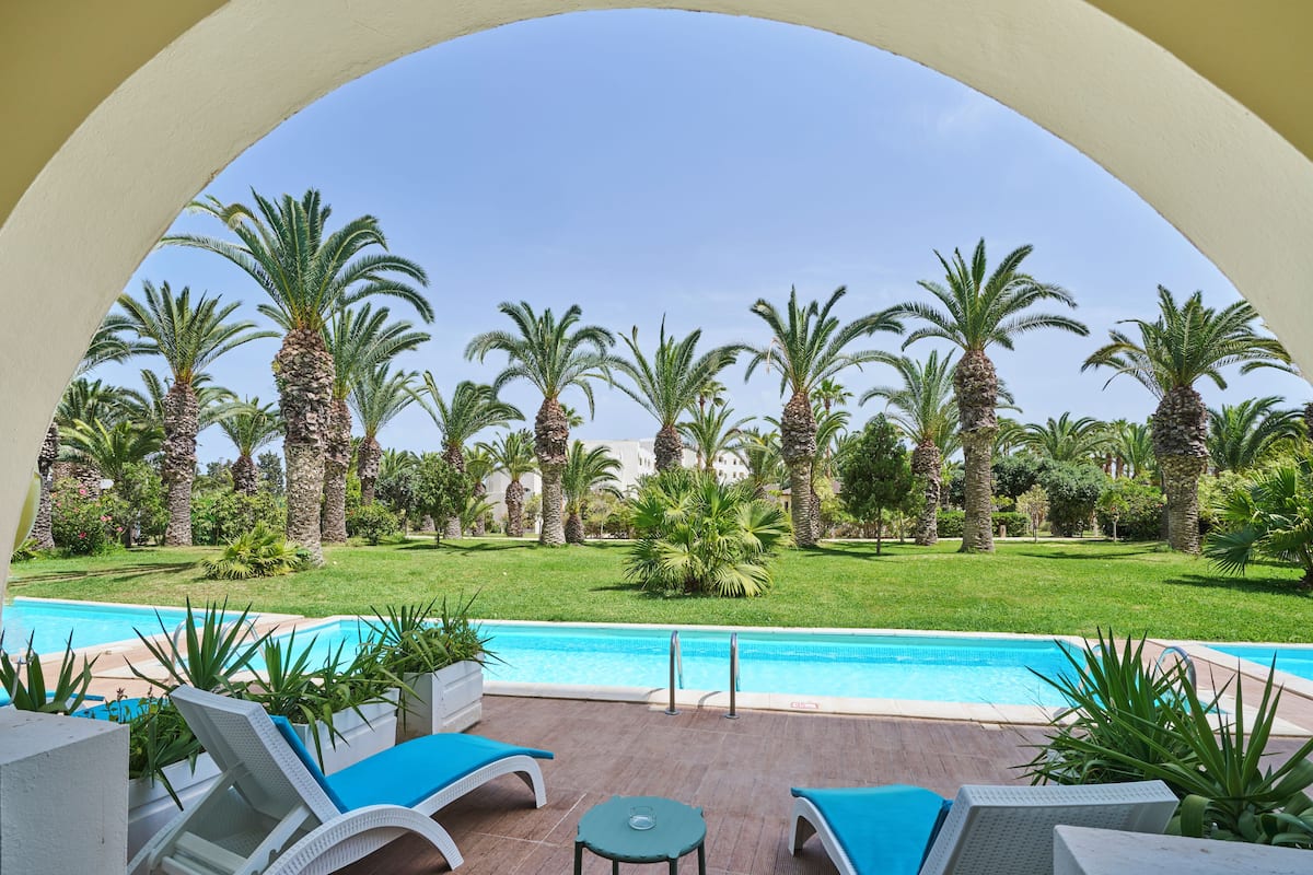 Un área junto a la piscina con sillones reclinables y una mesa, rodeada de palmeras y un cielo azul despejado.