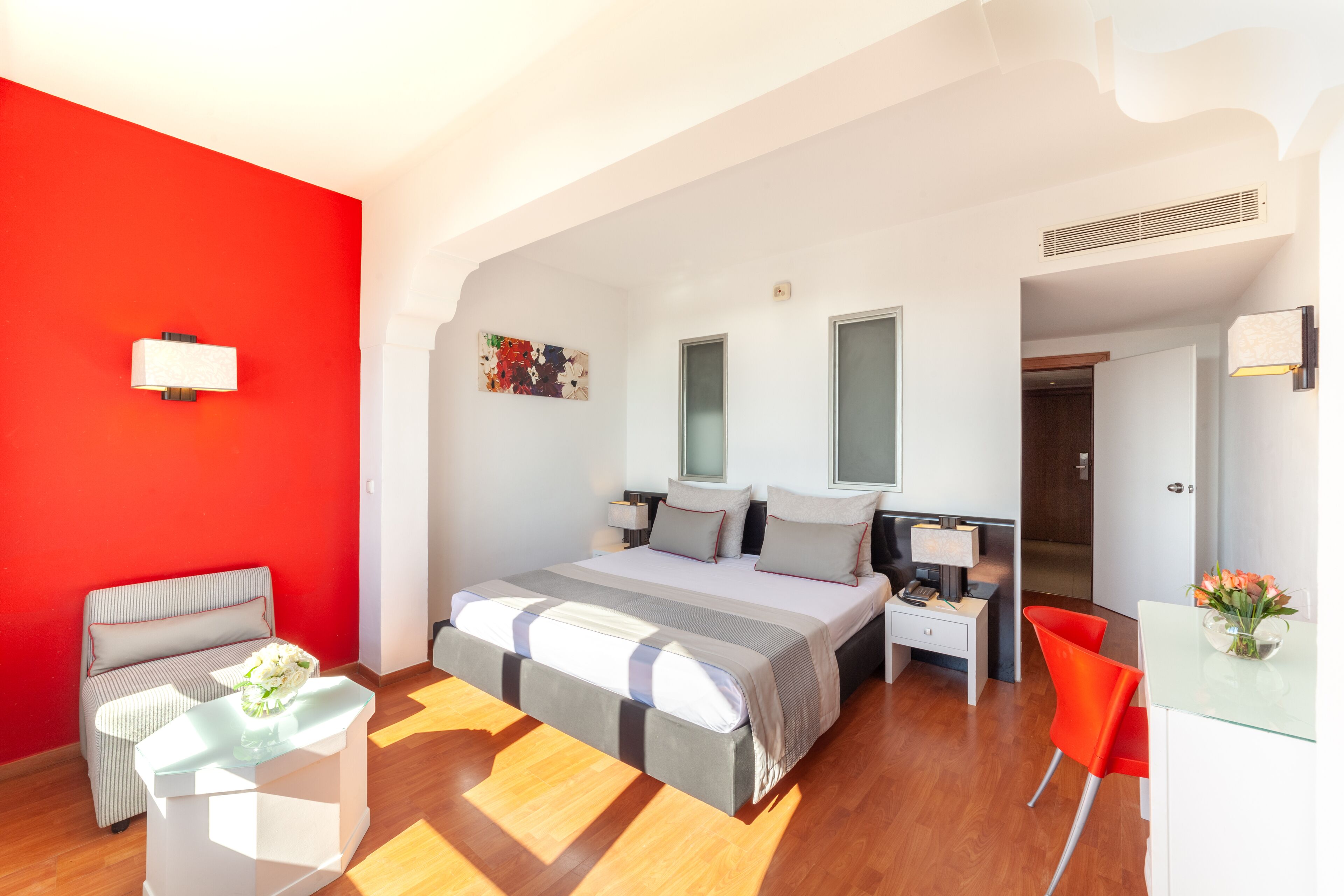 A modern hotel room with a large bed, a desk, a chair, and a red accent wall.