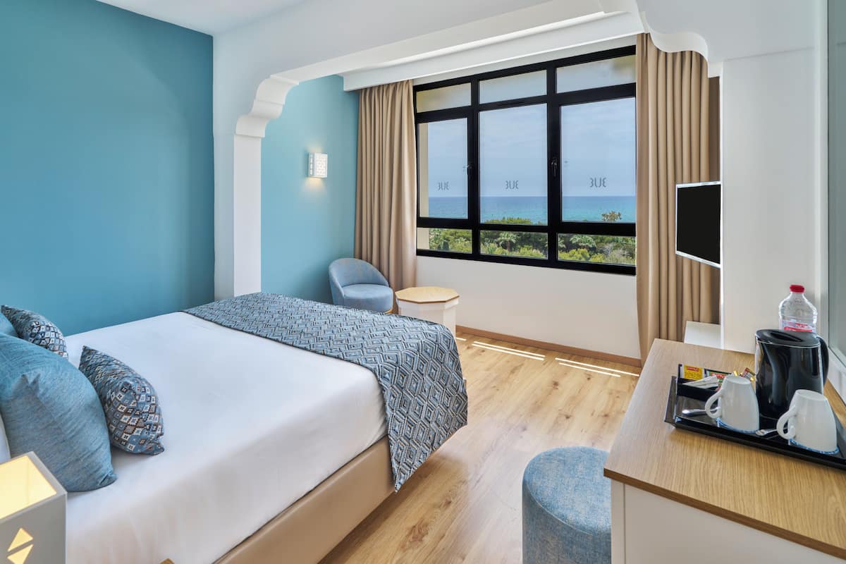 Habitación de hotel con cama, escritorio con bandeja de tazas de café, silla, televisor y ventana con vista al mar.