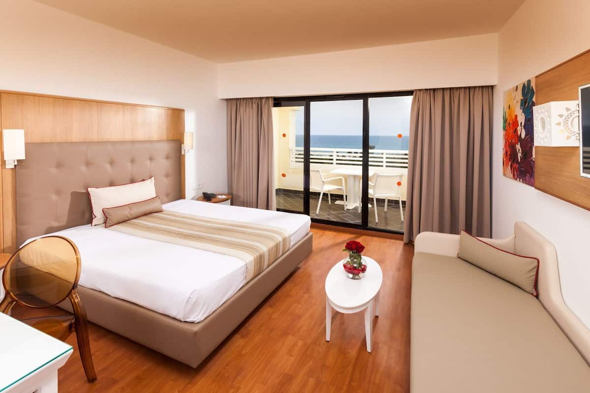 Una habitación de hotel con cama, sofá, una mesita con un florero, una silla y un balcón con vista al mar.