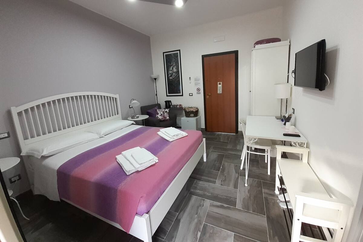 A modern hotel room with a bed, a desk, a chair, a TV, and a door.