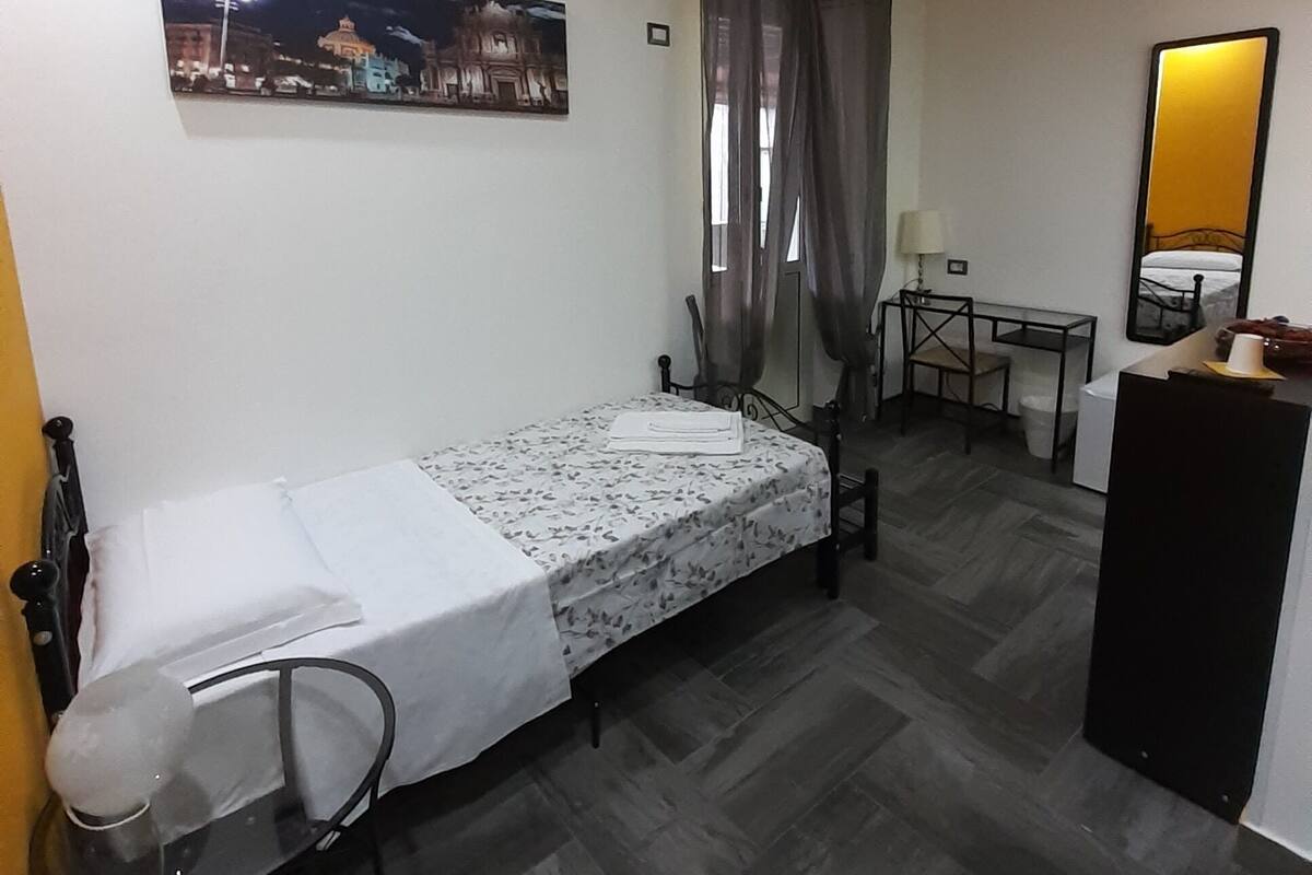 A compact hotel room with a single bed, a small desk, a chair, a mirror, and a picture of a cityscape.