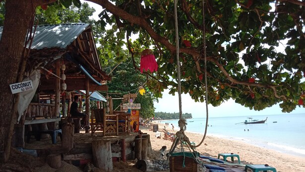 A beach scene with a wooden structure labeled "Coconut 60 Bath" and a sign for "Coca Cola." There are beach chairs and a hammock under a tree, and the ocean is visible in the background. - The Sea At Lanta Hotel (Ko Lanta)