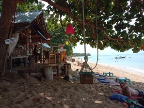 A beach setting with a wooden structure labeled 'Coconut 60 Bath' and a sign for 'Coca Cola.' There are beach chairs and a hammock under a tree. - The Sea At Lanta Hotel (Ko Lanta)
