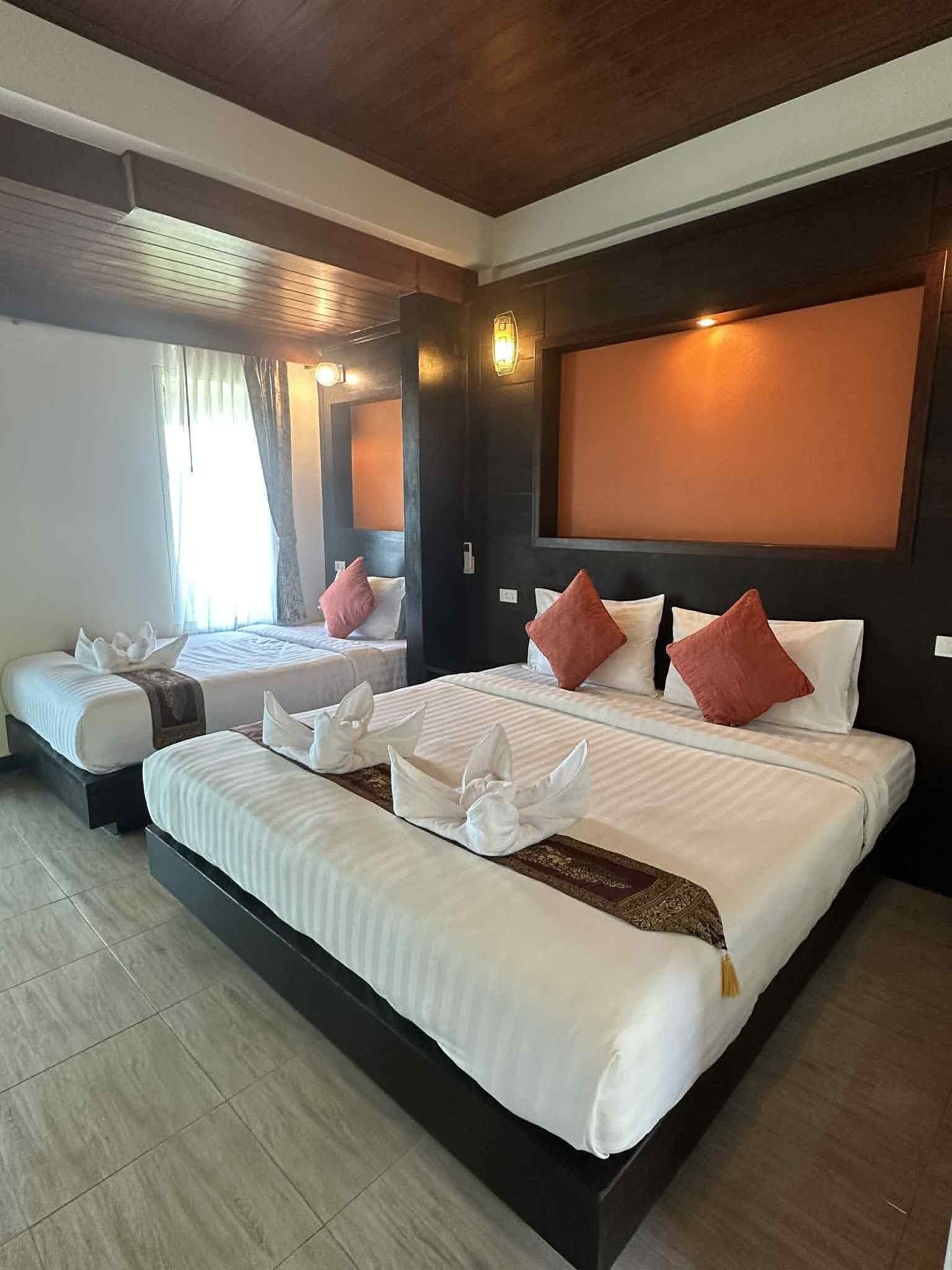 Quarto de hotel com duas camas, cada uma com lençóis brancos e almofadas vermelhas. O quarto tem teto de madeira e uma grande janela com cortinas.