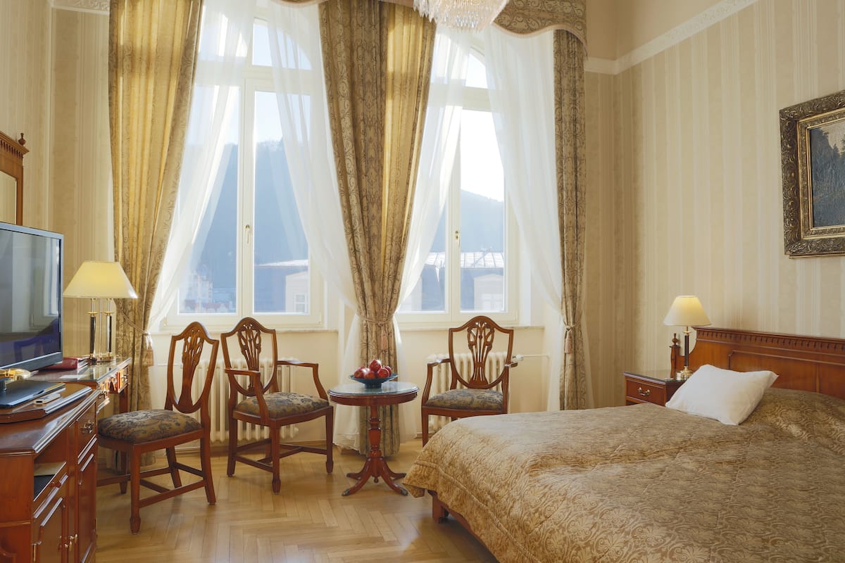 Ein Hotelzimmer mit einem großen Bett, zwei Holzstühlen, einem kleinen Tisch mit Obst, einem Fernseher und einem Gemälde an der Wand.