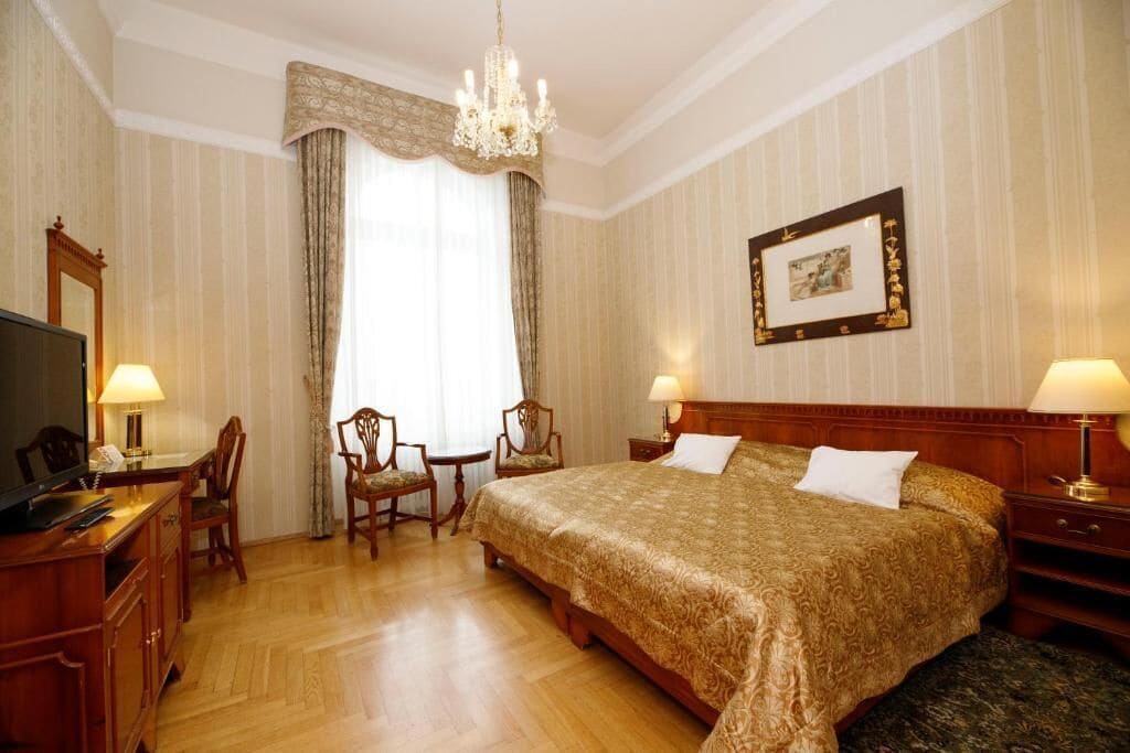 A hotel room with a large bed, a desk with a chair, a television, and a chandelier.