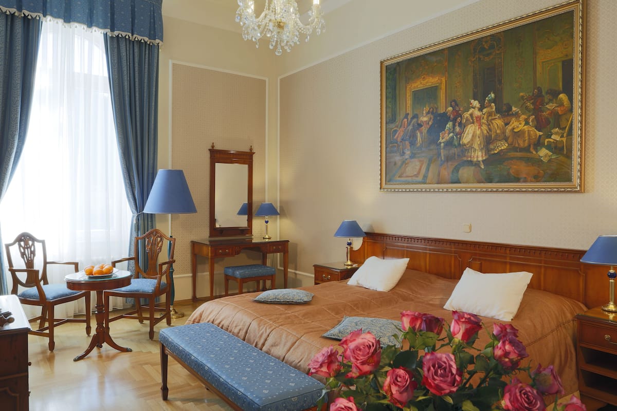 Ein Hotelzimmer mit einem großen Bett, zwei Nachttischen mit Lampen, einem Holztisch mit Stuhl, einem runden Tisch mit einem Stuhl und einem großen Gemälde an der Wand.