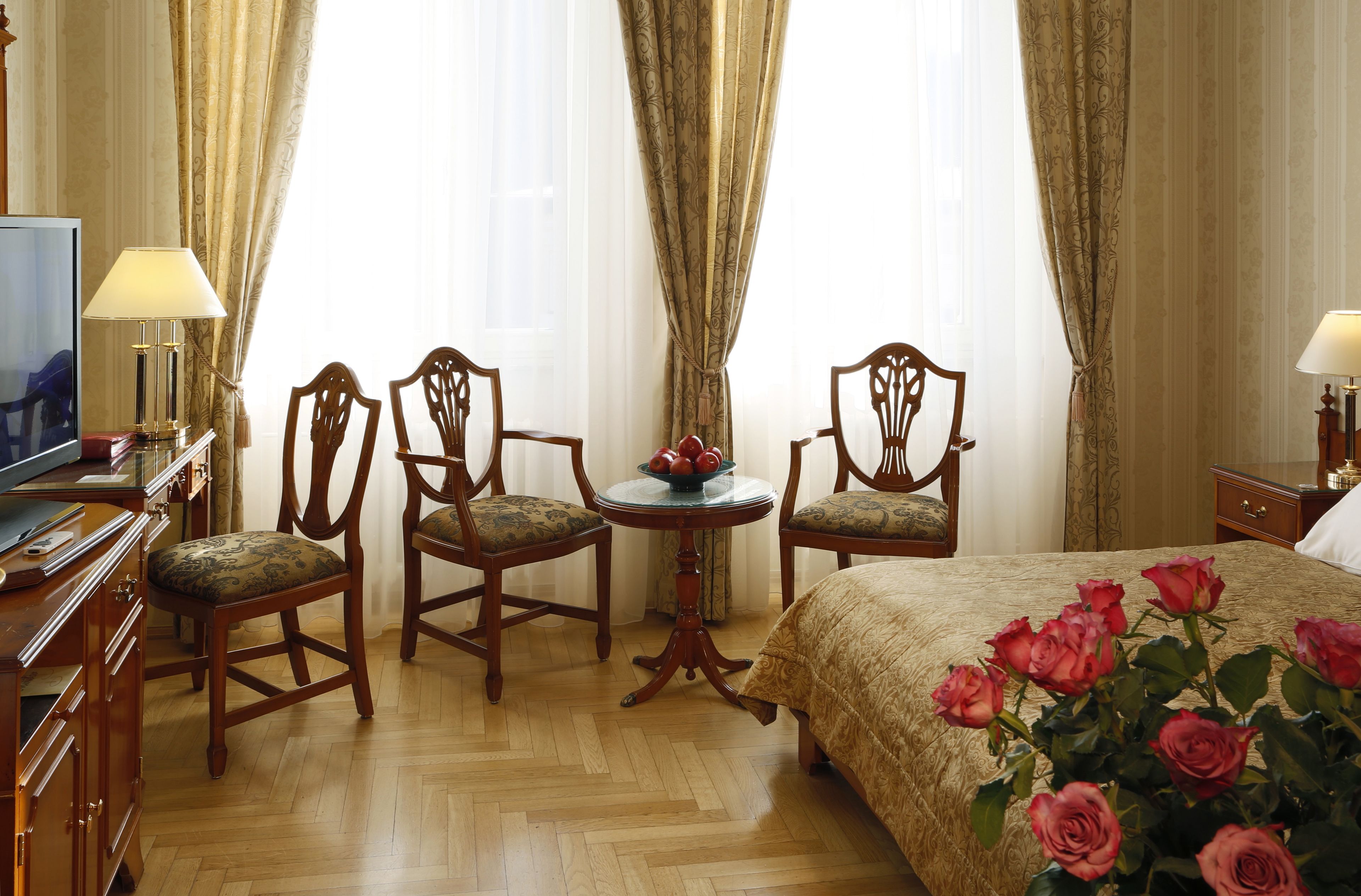 A hotel room with a wooden floor, a bed with a floral bedspread, two wooden chairs around a small round table, and a television on a wooden dresser.