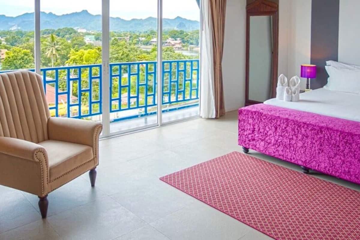 Quarto de hotel com uma cama, uma cadeira, varanda com grade azul e vista para o verde e montanhas.
