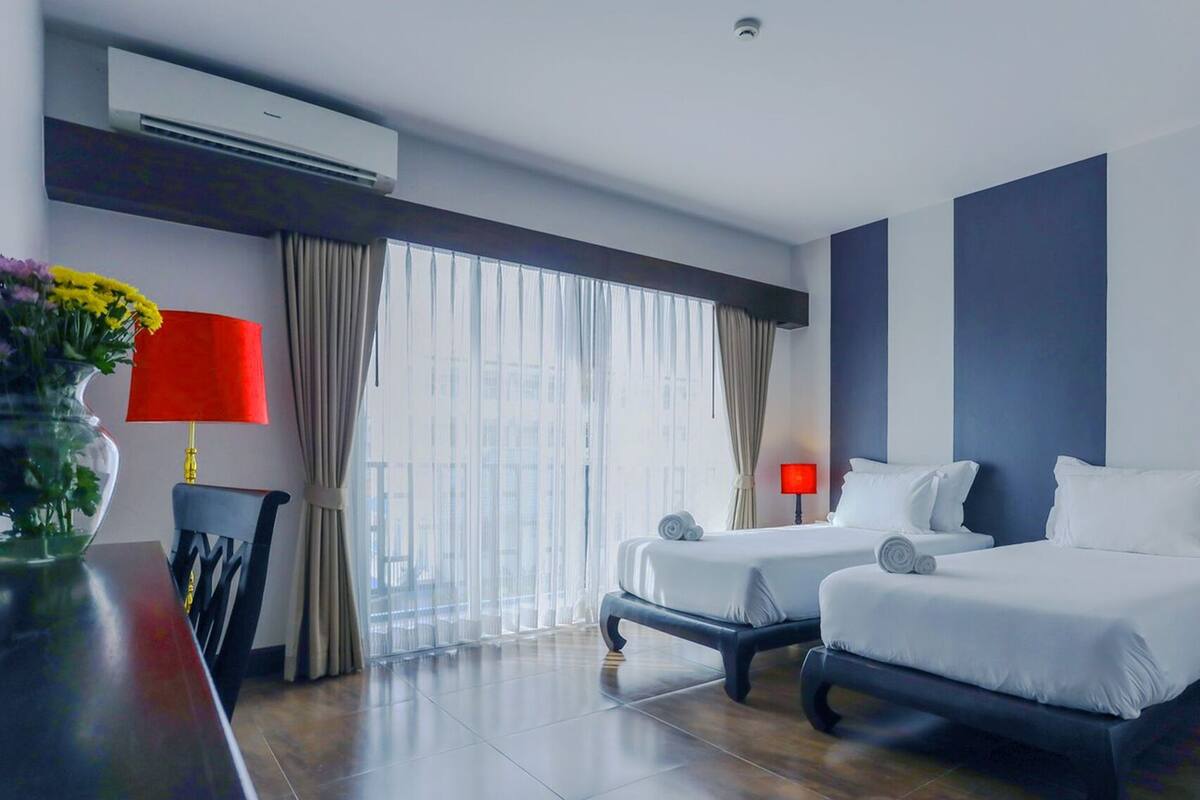 Quarto de hotel com duas camas, uma janela grande com cortinas, um abajur vermelho e um aparelho de ar condicionado.