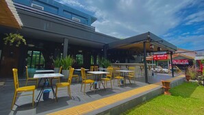Un espacio con mesas y sillas amarillas para disfrutar al aire libre, una zona cubierta y un edificio con fachada azul.