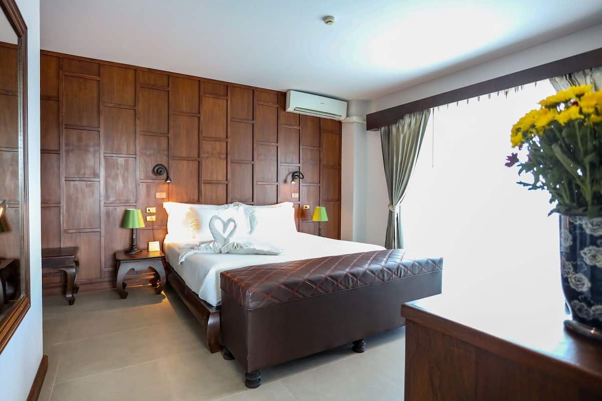 Quarto de hotel com parede de painel de madeira, cama com travesseiro em formato de coração, duas luminárias de cabeceira, uma janela grande com cortinas e um vaso de flores.