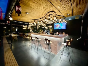 A modern bar area with a high ceiling, exposed wooden beams, and a central bar counter with stools. - The Bridge Residence Hotel (Kanchanaburi)