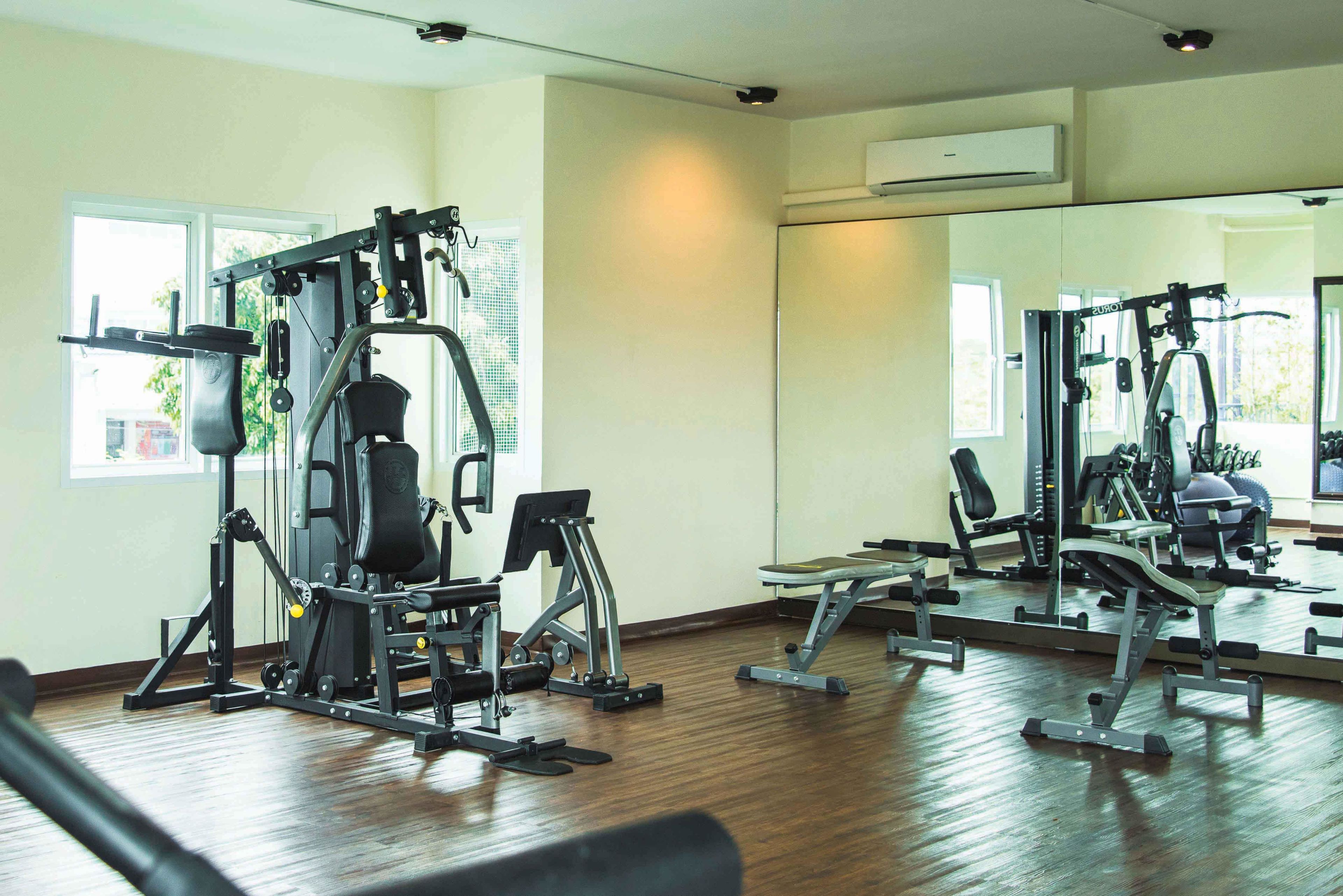 Ein Fitnessstudio mit verschiedenen Trainingsgeräten, großen Fenstern und einem Spiegel, der die Geräte reflektiert.