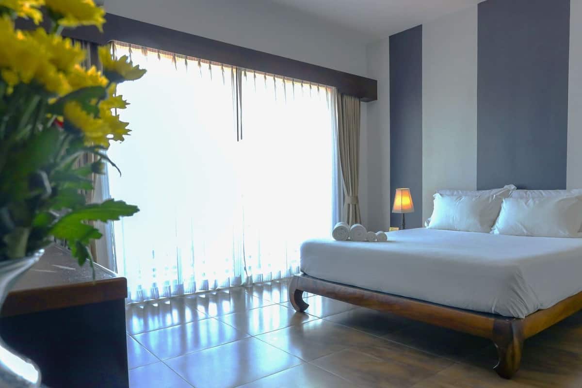 Cama bem arrumada com roupa branca, duas luminárias de cabeceira e uma planta em vaso à esquerda.