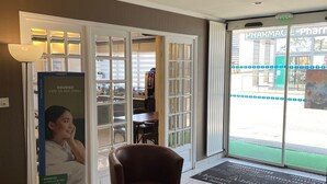 Een hotel lobby met een receptiebalie, een wachtruimte met een bruine stoel, een poster die het hotel promoot en een uitzicht op een apotheek door de glazen deuren.
