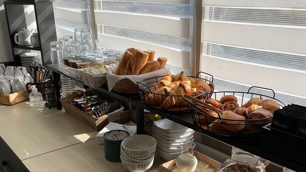 Een buffetopstelling met diverse etenswaren en bestek, waaronder brood, gebak en toevoegingen, gerangschikt op een toonbank met uitzicht naar buiten door grote ramen.