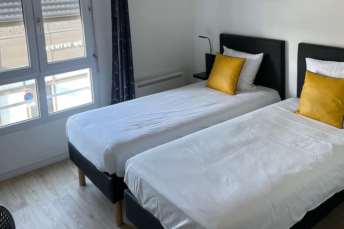 Une chambre d’hôtel avec deux lits, un luminaire au plafond, un tableau et une fenêtre avec des stores.