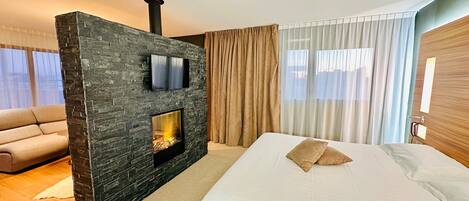 A modern hotel room with a stone fireplace, a flat-screen TV, a large bed, and a view of the city through sheer curtains.