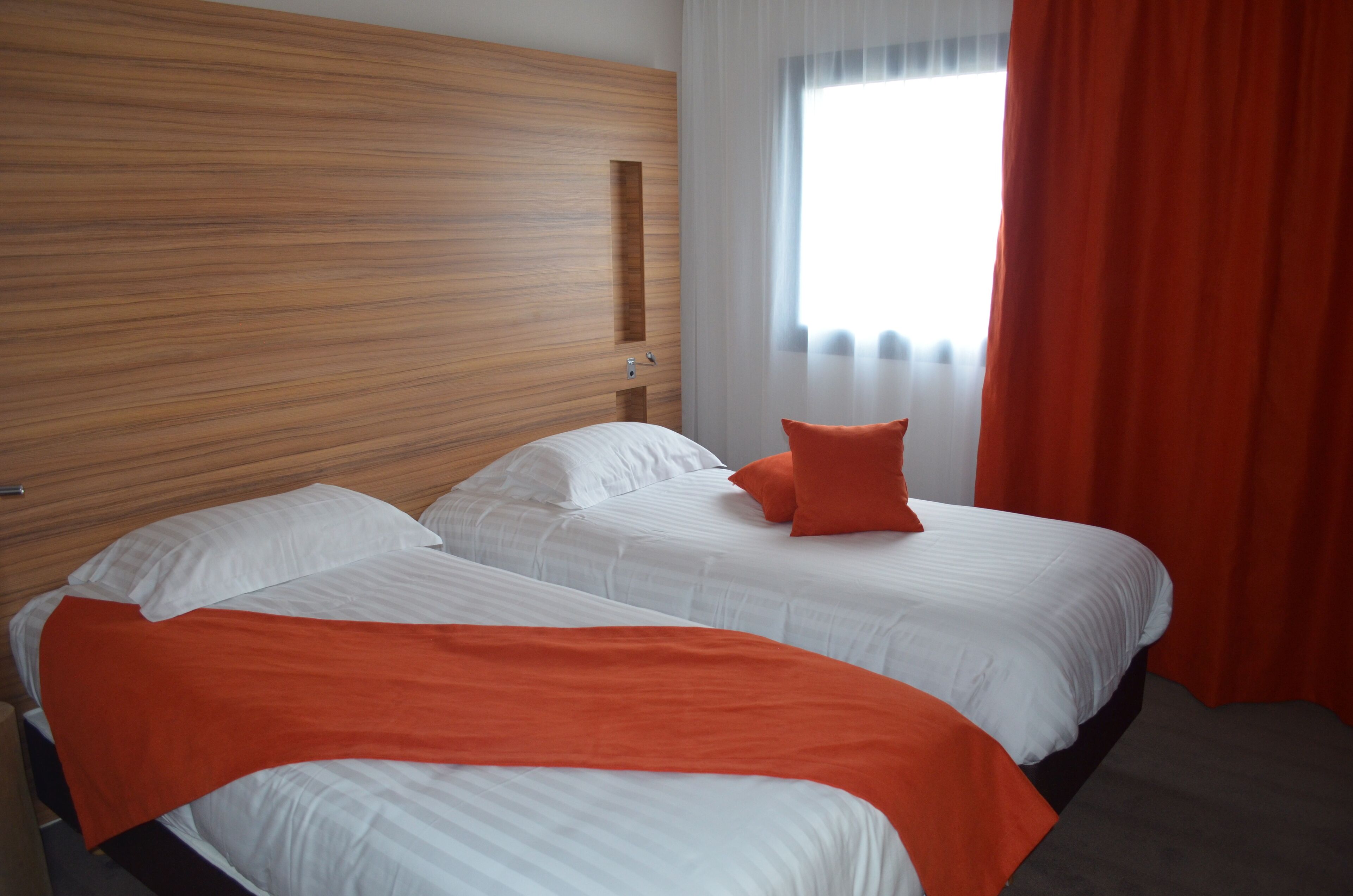 Une chambre d’hôtel avec deux lits, une tête de lit en bois, du linge de lit blanc et une fenêtre avec des rideaux légers.