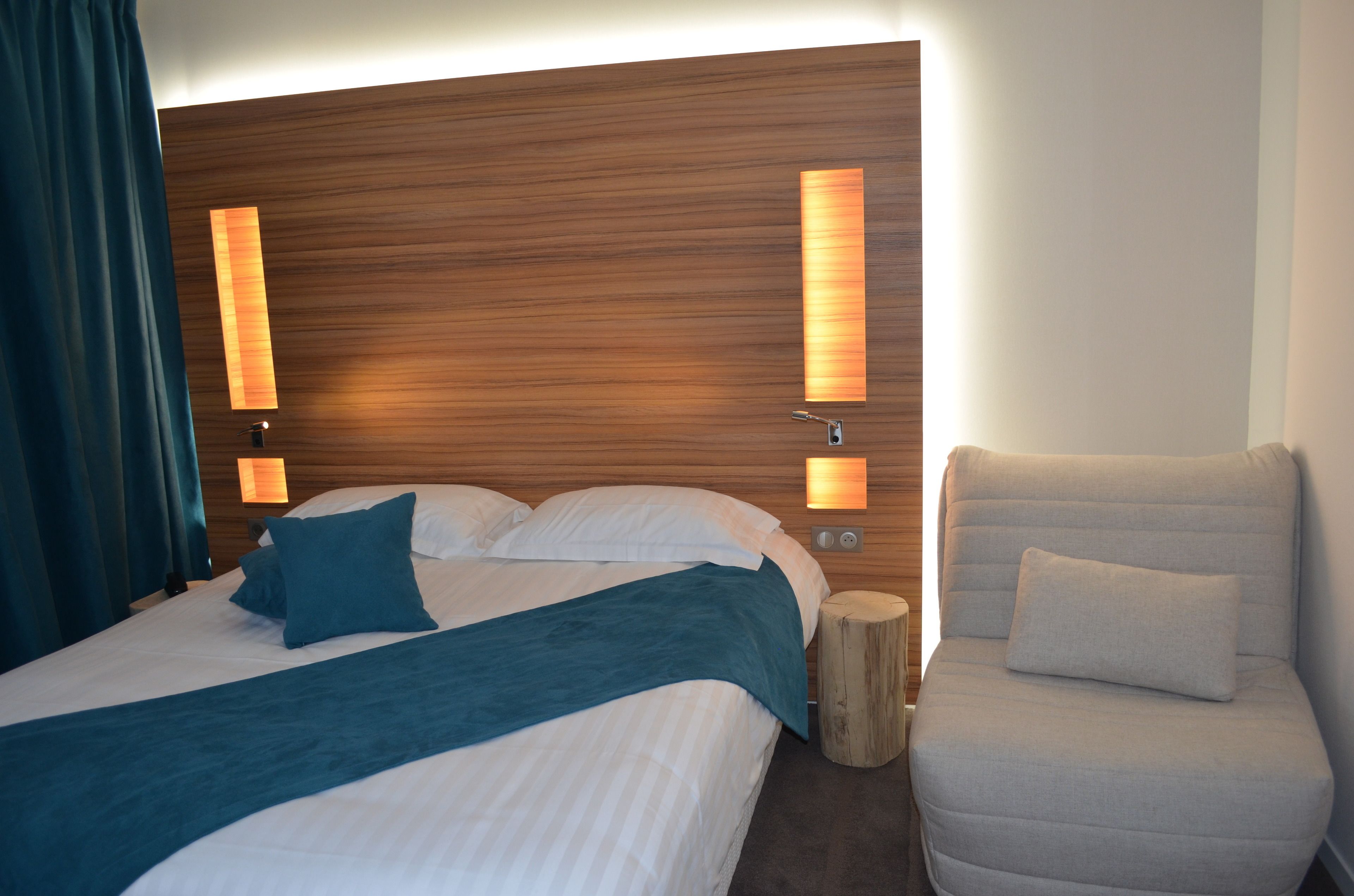 Une chambre d’hôtel avec un grand lit, une tête de lit en bois, deux lampes fixées au mur, une table de chevet et une chaise.