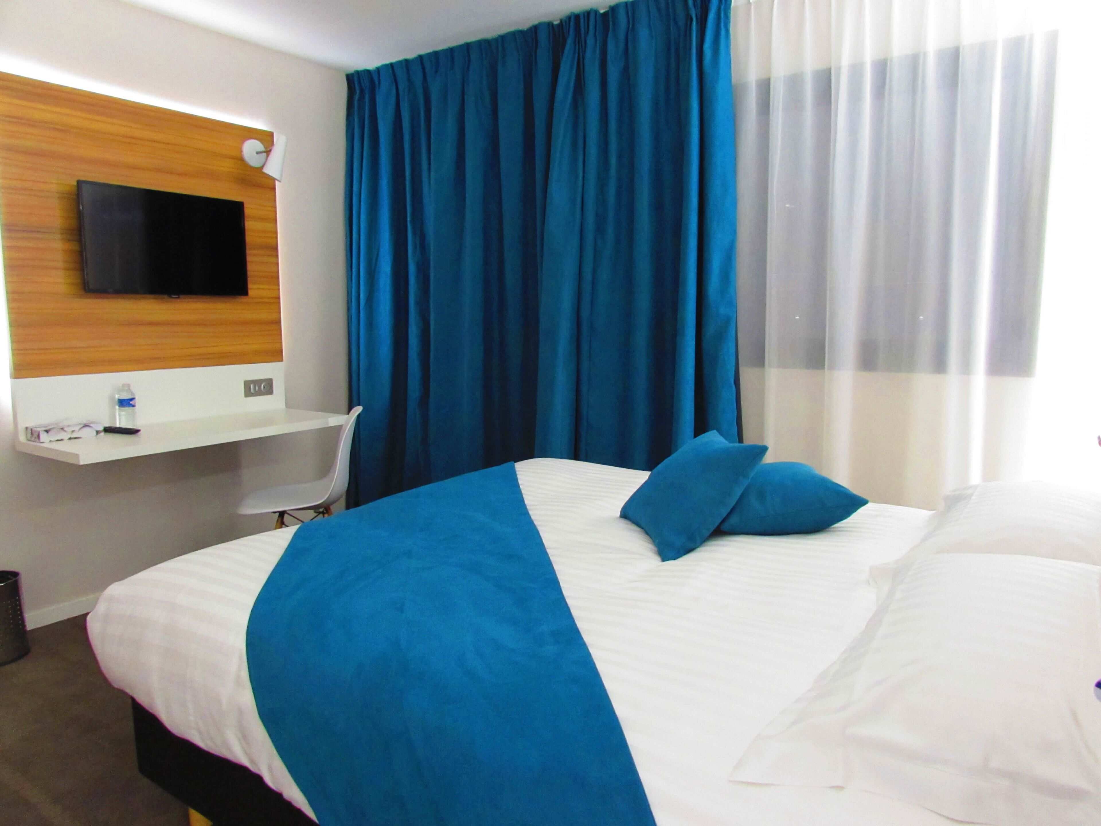 Ein Hotelzimmer mit Bett, Schreibtisch, Flachbildfernseher, Stuhl und Fenster mit blauen Vorhängen.