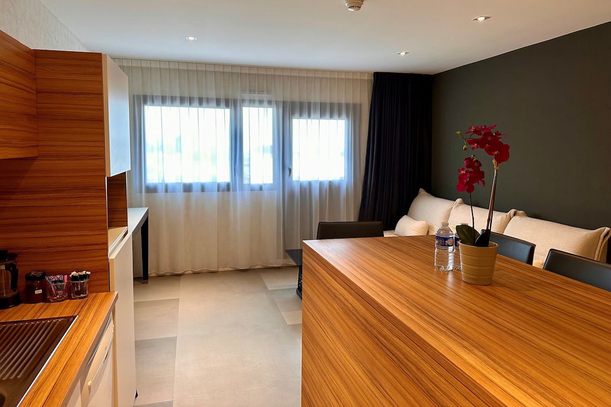 Ein modernes Hotelzimmer mit einem hölzernen Empfangstresen, einem Essbereich mit Holztisch, einer Couch und einer Topforchidee.