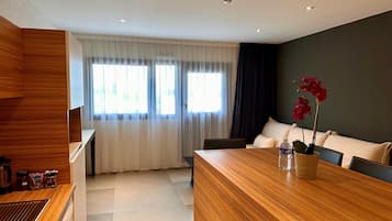 A modern hotel room with a wooden reception desk, a dining area with a wooden table, a sofa, and a potted red orchid.