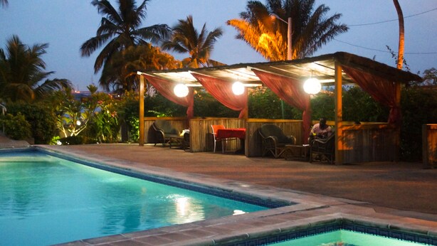 A swimming pool with a covered seating area, palm trees, and a wooden fence.